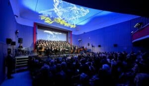 Široki kadar dvorane s publikom i zborom pod plavim svjetlima tijekom izvedbe projekta "The Armed Man" 2026. godine.