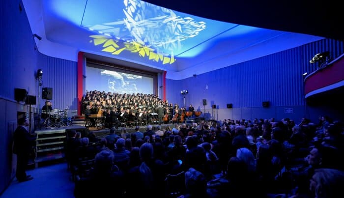 Široki kadar dvorane s publikom i zborom pod plavim svjetlima tijekom izvedbe projekta "The Armed Man" 2026. godine.