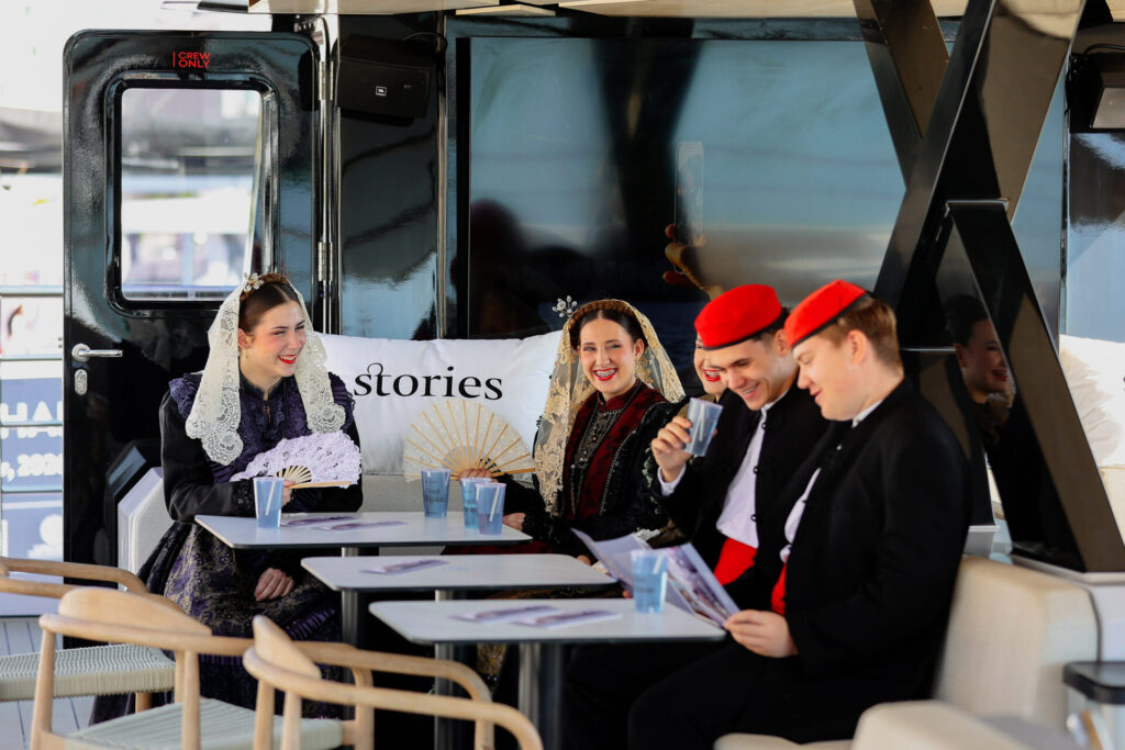 Mladi u tradicijskim nošnjama sjede za stolom s natpisom Stories na platformi Stories Croatia tijekom sajma u Splitu Izvor: HGK.